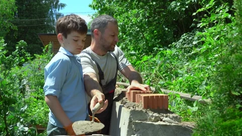 Dad teaching son to put up brick wall. Joyful young family working in the garden Stock Footage 234785694