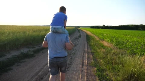 Daddy goes with small son sitting on shoulders through rural road near meadows Stock Footage 304111666