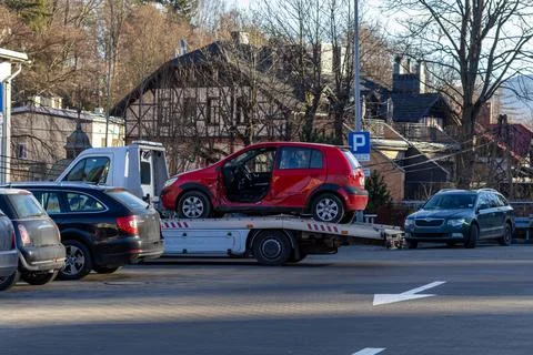 A damaged red hatchback car rests on a tow truck bed parked on an urban street, Fotos Stock