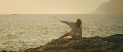 Dancer bends low on rocky shore during sunset in a moment of improvisation Video stock 319877240