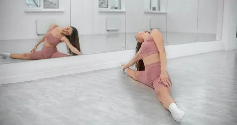 Dancer is sitting on the twine and doing bends in front of the mirror wall in Stock Footage 145676448