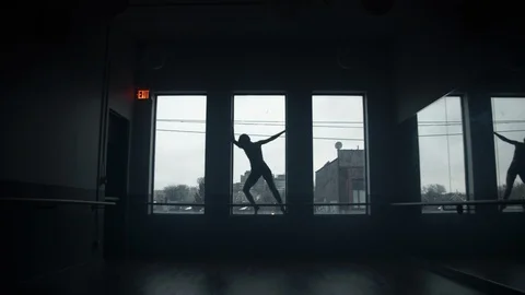Dancer on window sill dancing, leaning back in large dark dance studio 스톡 동영상 104275535