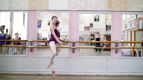In dancing hall, Young ballerina in purple leotard performs developpe attitude Stock Footage 89013262
