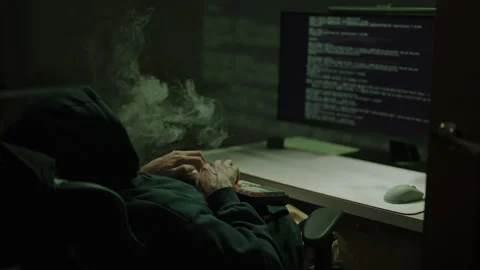 Dangerous hooded hacker typing code sitting at desk, vape smoke curling around Stock Footage 325715229
