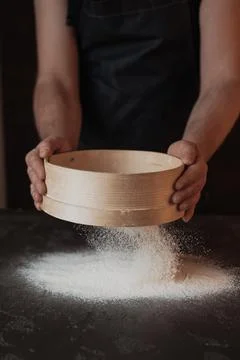 A dark background baker's hands sift white wheat flour over a sieve over Stock Photos