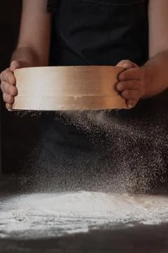 A dark background baker's hands sift white wheat flour over a sieve over Stock Photos