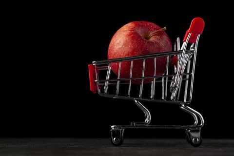 Dark background image with a toy shopping cart filled with a red apple A conc Stock Photos