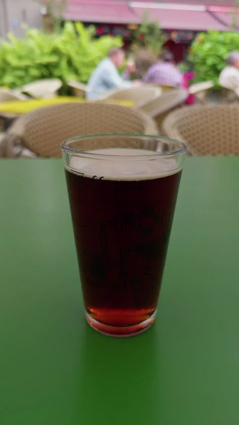 Dark beer in clear pint glass on green table with blurred outdoor cafe seating Stock Footage 338134456