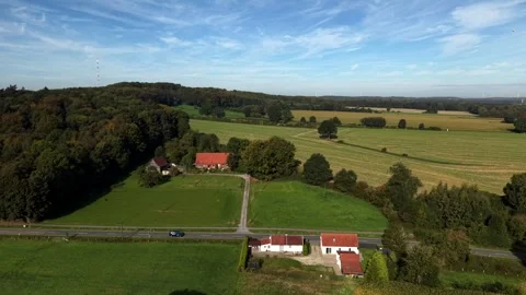 Dark car driving along rural road past farm houses with red tile roofs, green Stock Footage 346797953