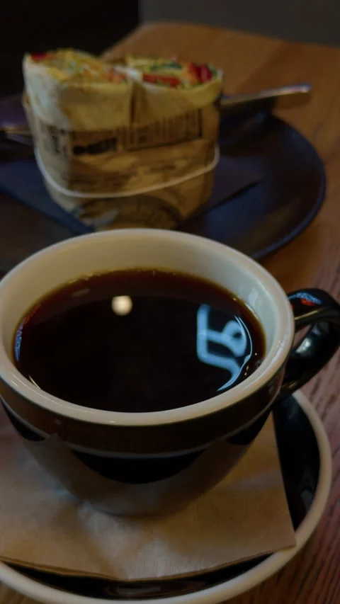 Dark coffee cup on table with wrapped sandwich in soft focus cafe meal setting Stock Footage 345605289