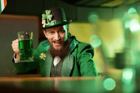 Dark-haired bearded young man in a leprechaun hat with shamrock looking funny Stock Photos