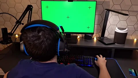 A dark- haired boy uses a computer, sits with headphones in front of a green 스톡 동영상 271082033