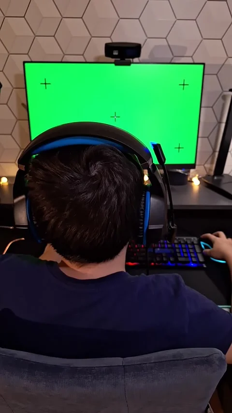 A dark- haired boy uses a computer, sits with headphones in front of a green 스톡 동영상 276192875