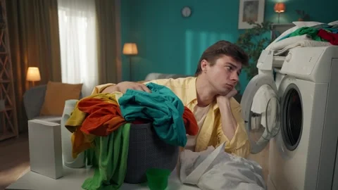Dark-haired man sitting next to a washing machine, looking at the laundry basket Stock Footage 252387344