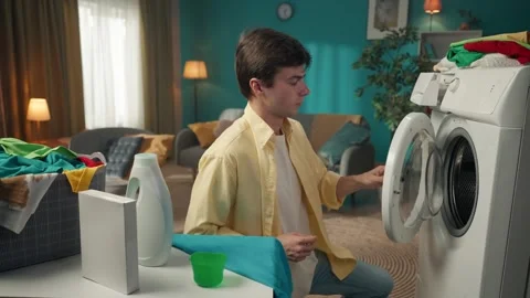 Dark-haired man sitting next to a washing machine, pouring a liquid detergent Stock Footage 252388506