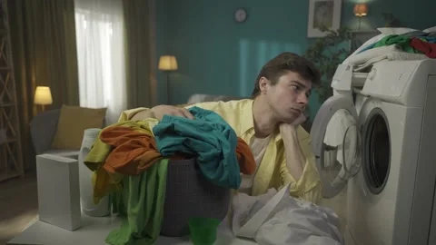Dark-haired man sitting next to a washing machine, looking at the laundry basket Stock Footage 252390339
