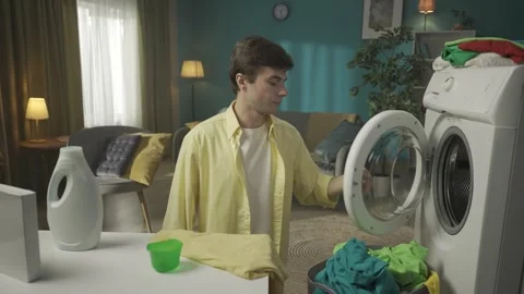 Dark-haired man sitting next to a washing machine, pouring a liquid detergent Stock Footage 252391864