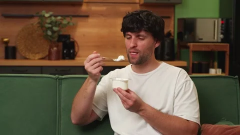A dark-haired man in a white T-shirt eats delicious yogurt sitting on a green Stock-Footage 249682029