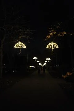A dark park pathway is illuminated by multiple glowing umbrella lights Stock Photos