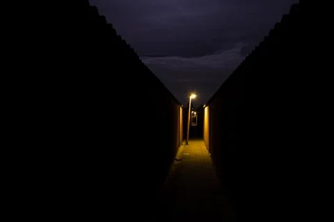 A dark path between houses on a cloudy night. A streetlights illuminate the path Stock Photos