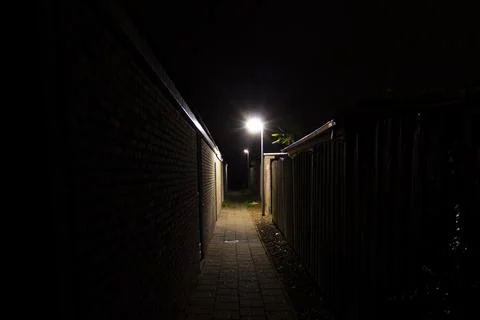 A dark path between houses at night. A single streetlight illuminates the path Stock Photos