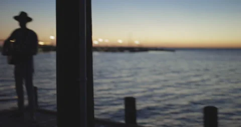 Dark silhouette of man with hat walking on pier and playing guitar after sunset 스톡 동영상 147063928