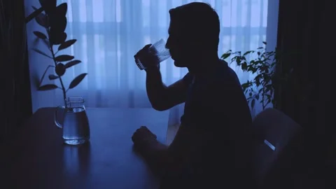 Dark silhouette of man sitting at table near window drinking glass of water Stock Footage 314780264