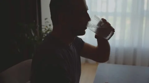 Dark silhouette of man sitting at table near window drinking glass of water Stock Footage 314780411