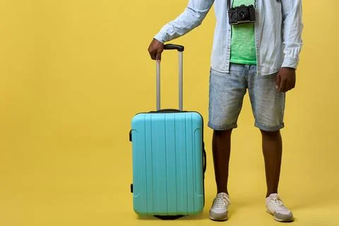 Dark-skinned guy in modern summer clothes is holding tourist suitcase by handle Stock Photos