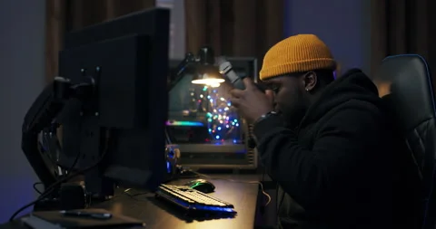 Dark-skinned guy in a yellow cap sits at desk getting ready to play a video game Vídeo Stock 178738803