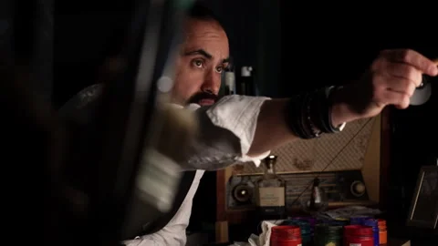 A dark-skinned man with a beard in a shirt and vest sits at a table and paints Stock Footage 241493917