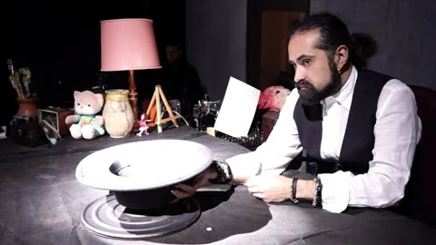 A dark-skinned man with a beard in a shirt and vest sits at a table, takes off Stock-Footage 241494750