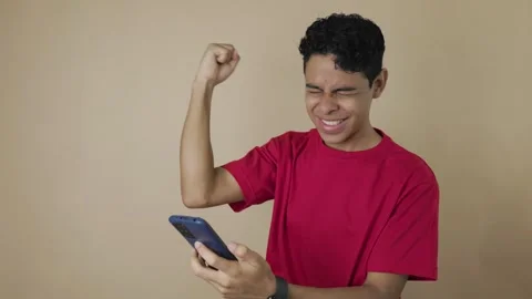 Dark skinned man celebrating with his cell phone on a light brown background Stock Footage 315062017