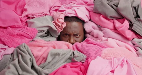Dark skinned man covered with heap of clothes cluttered and tries to clean out Stock Footage 157679599