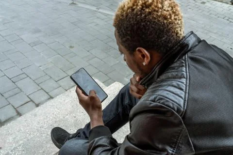 Dark-skinned man looking at mobile phone screen and sitting on concrete steps. Stock Photos