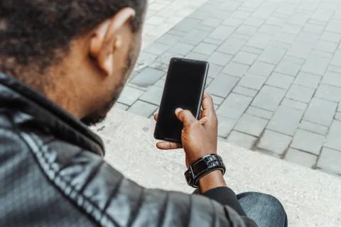 Dark-skinned man looking at mobile phone screen and sitting on concrete steps. Stock Photos