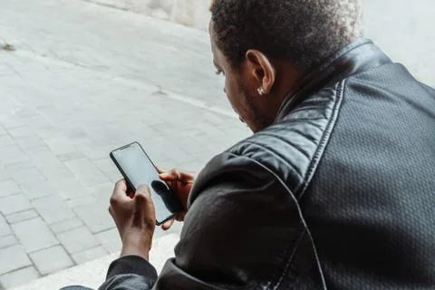 Dark-skinned man looking at mobile phone screen and sitting on concrete steps. Stock Photos