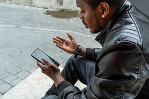 Dark-skinned man looking at mobile phone screen and sitting on concrete steps. Stock Photos