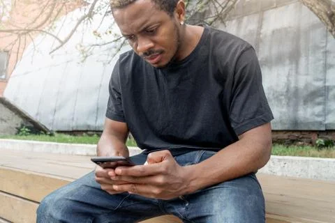 Dark-skinned man looking at mobile phone screen while sitting under tree Stock Photos
