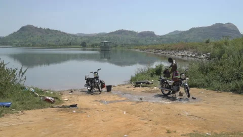Dark skinned man washes his motorcycle by the lake Stock Footage 100637738