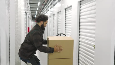 Dark-skinned man in work overalls putting boxes on cart in self storage facility Video stock 259435729
