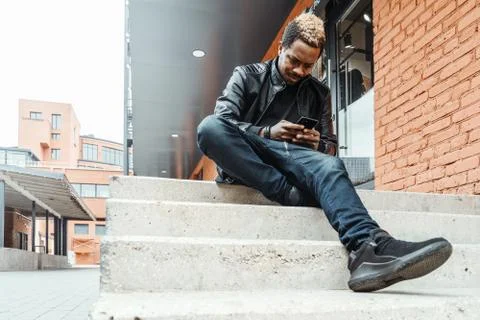Dark-skinned smiling man looking at mobile phone screen and sitting on steps. Stock Photos