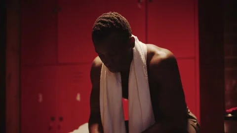 Dark-skinned young man sits in a locker room with a towel around his neck Stock Footage 146552594