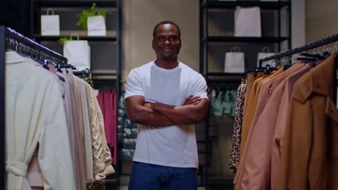 A dark-skinned young man in a white T-shirt stands btw rails with clothes and Stockbeeldmateriaal 267647013
