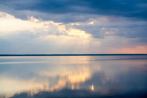 Dark storm clouds and soft sunset light above peaceful lake with reflections  Stock Photos