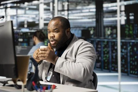 Data center technician drinking coffee analyzing software logs metrics on PC Stock Photos