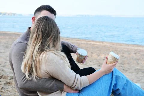 Date on the beach. A young couple in sweaters sits on a plaid, holds coffee i Stock Photos