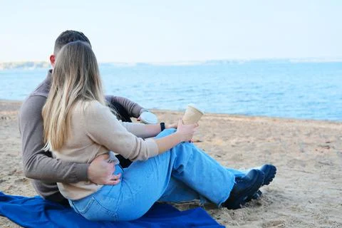 Date on the beach. A young couple in sweaters sits on a plaid, holds coffee i Stock Photos