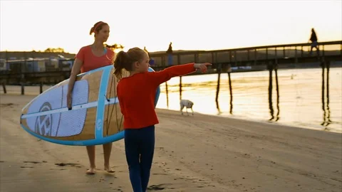 Daughter points out to sea while her mother carries paddleboard. Vidéo 88369335