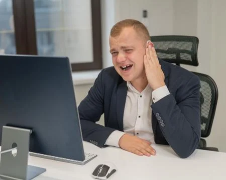A deaf man works at a computer in the office. Broken hearing aid causing pain. Stock Photos
